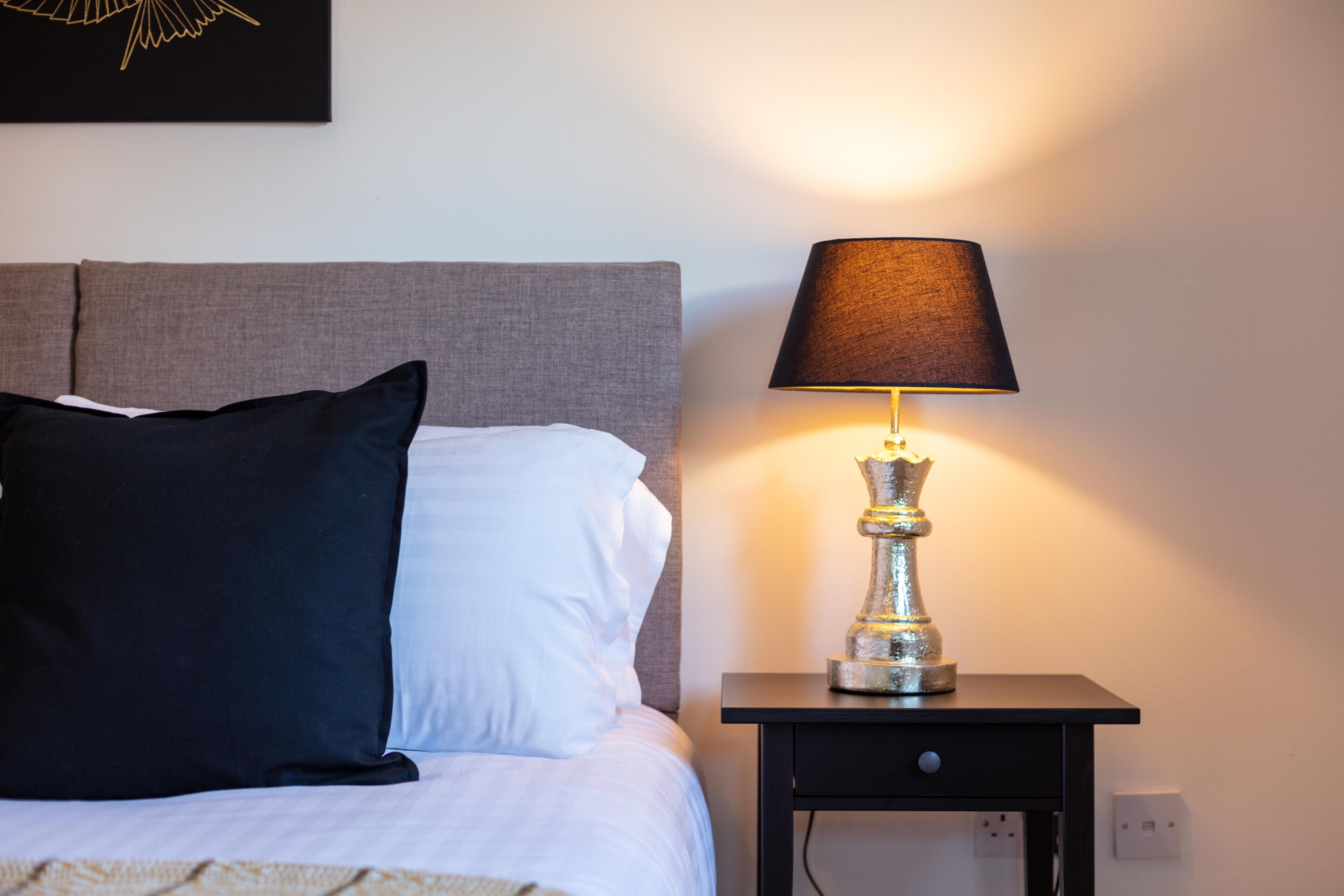 Una cama bien hecha con almohadas blancas y negras, un cabecero gris y una lámpara decorativa en una mesita de noche.