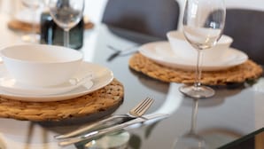 Een eettafel gedekt met witte borden, een wijnglas en bestek op geweven placemats.