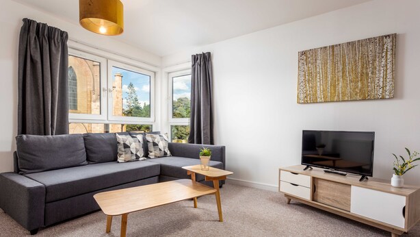 Een moderne woonkamer met een grijze bank, een houten salontafel, een televisie op een houten kast en een groot raam met gordijnen.