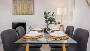 Een eethoek met een glazen tafel, grijs gestoffeerde stoelen, witte borden en een vaas met groene bladeren.