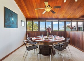 A dining area with a round table, wicker chairs, a ceiling fan, and a view of the ocean through large windows.