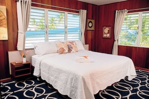 A neatly made bed with a white bedspread and decorative pillows, a bedside table with a vase of flowers, and a window with curtains. - Vacation Rental Redefined, St. George's Caye Belize (Belize City)