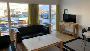 A modern living room with a black leather sofa, a wooden coffee table, and a TV on a wooden desk. Large windows offer a view of a snowy cityscape. - Nuuk Hotel Apartments by HHE (Nuuk)