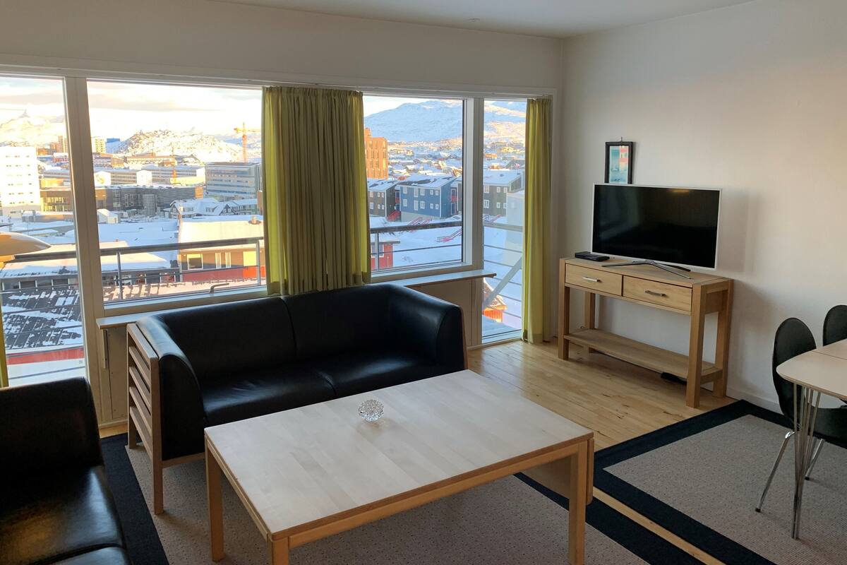 A modern living room with a black leather sofa, a wooden coffee table, and a TV on a wooden desk. Large windows offer a view of a snowy cityscape.