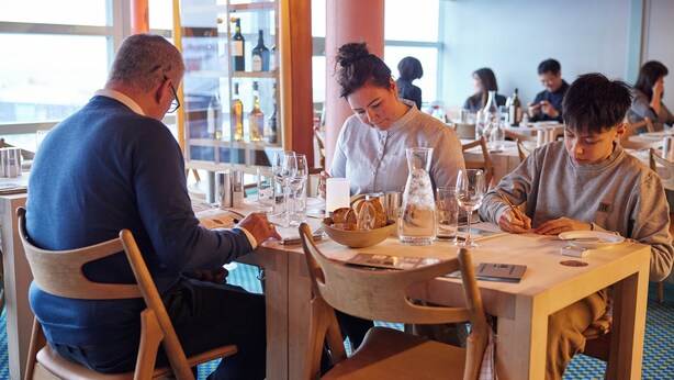A restaurant with wooden tables and chairs, a blue patterned floor, and a view of the sea through large windows. - Nuuk Hotel Apartments by HHE (Nuuk)