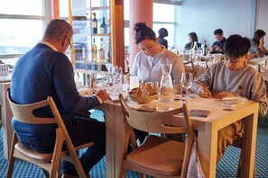 A restaurant with wooden tables and chairs, a blue patterned floor, and a view of the sea through large windows. - Nuuk Hotel Apartments by HHE (Nuuk)