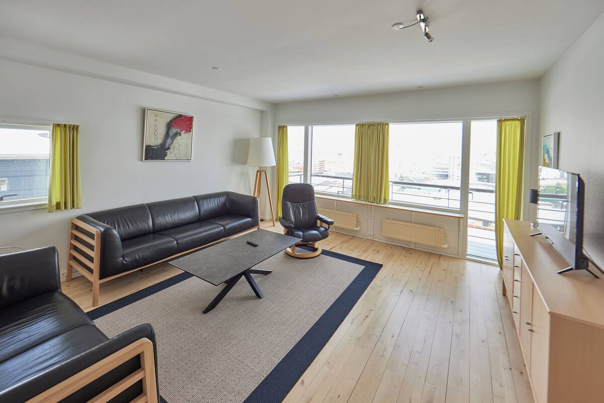 A modern living room with a black leather sofa, a wooden coffee table, a black armchair, and a TV on a wooden cabinet.