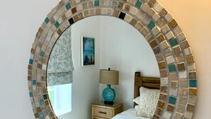 A round mirror with a mosaic frame, a bed with white bedding, a nightstand with a lamp, and a painting on the wall. - Beautiful Beachfront Apartment: Turtles Nest B (Dover)