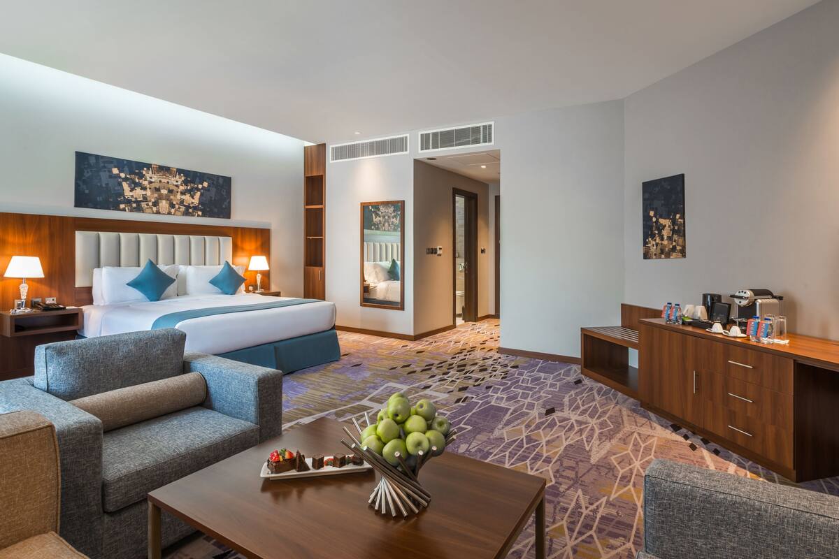 Une chambre d’hôtel comprenant un lit, deux tables de chevet avec des lampes, un canapé, une table basse agrémentée d’un arrangement de fruits et un bureau avec un ordinateur et d’autres objets.