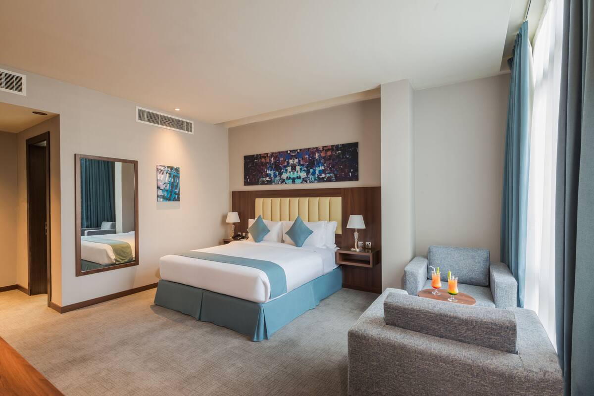 Une chambre d’hôtel avec un grand lit, un coin salon, un tableau au mur et une fenêtre avec des rideaux.