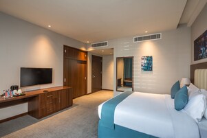 A hotel room with a bed, a desk with a flat-screen TV, a wardrobe, and a bathroom visible through an open door.