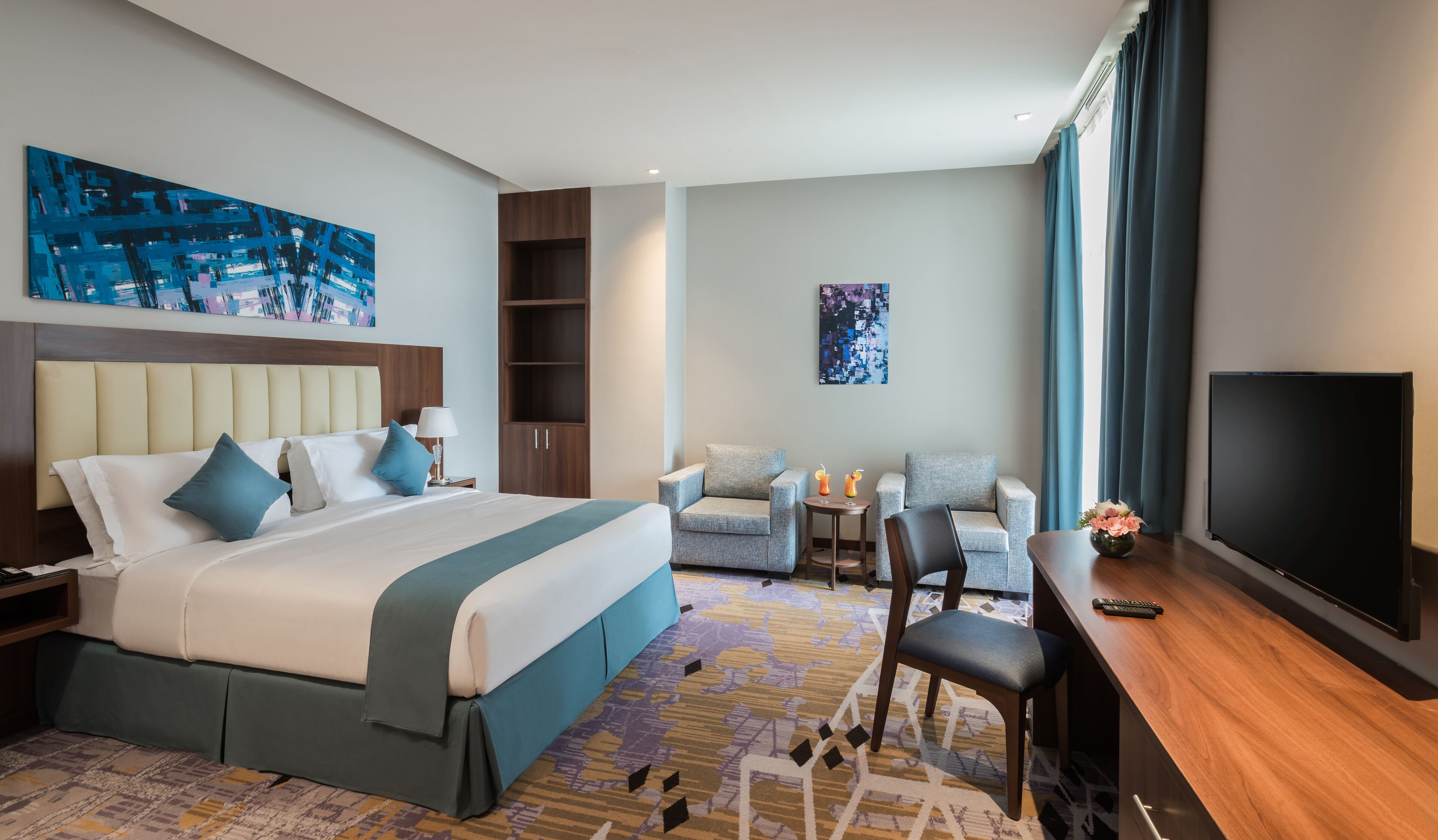 A hotel room with a large bed, two armchairs, a small table, a TV, and artwork on the walls.
