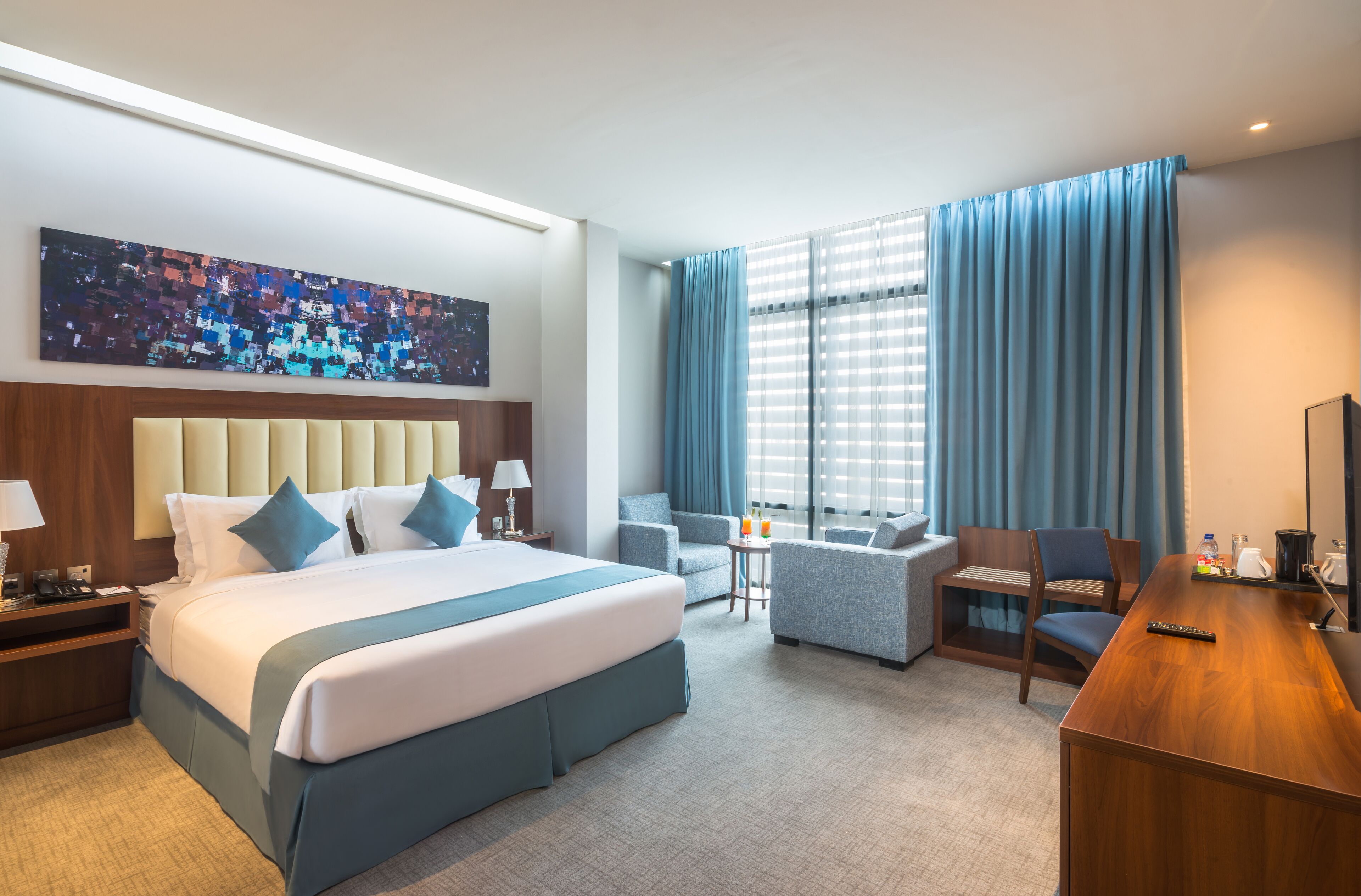 Quarto de hotel com uma cama grande, uma escrivaninha, um sofá e uma cadeira. Há uma janela com cortinas, um quadro na parede e um abajur na mesa de cabeceira.