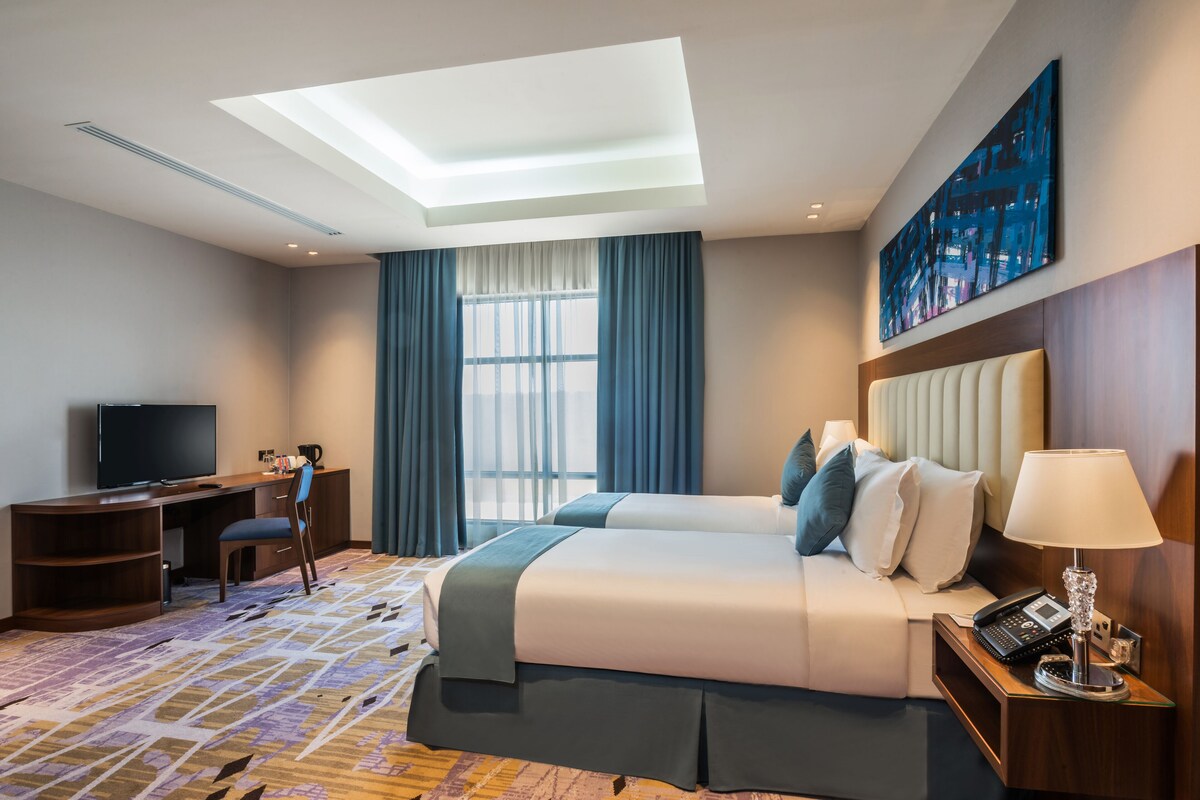 Una habitación de hotel con dos camas, un escritorio, un televisor y un ventanal con cortinas.
