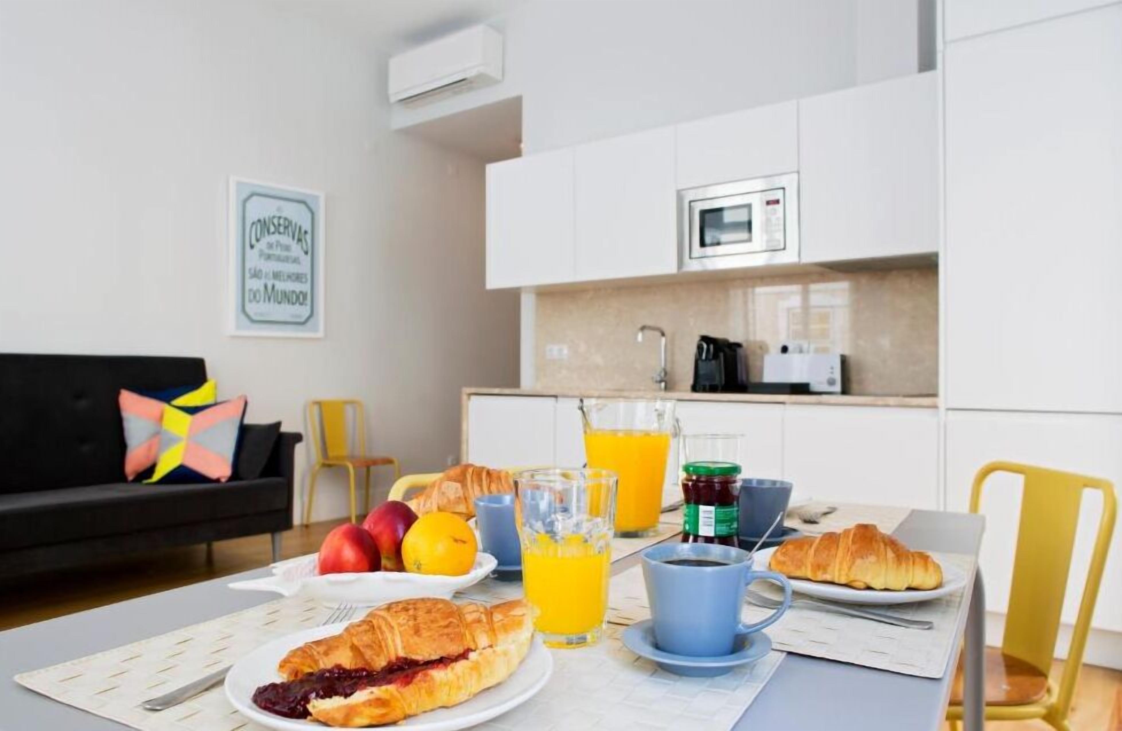 A breakfast setting with croissants, fruit, and orange juice on a table, with a kitchen in the background.