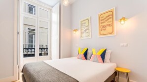 Ein Schlafzimmer mit einem Bett, einem kleinen Hocker, einem Fenster mit Aussicht und zwei Wandleuchten.