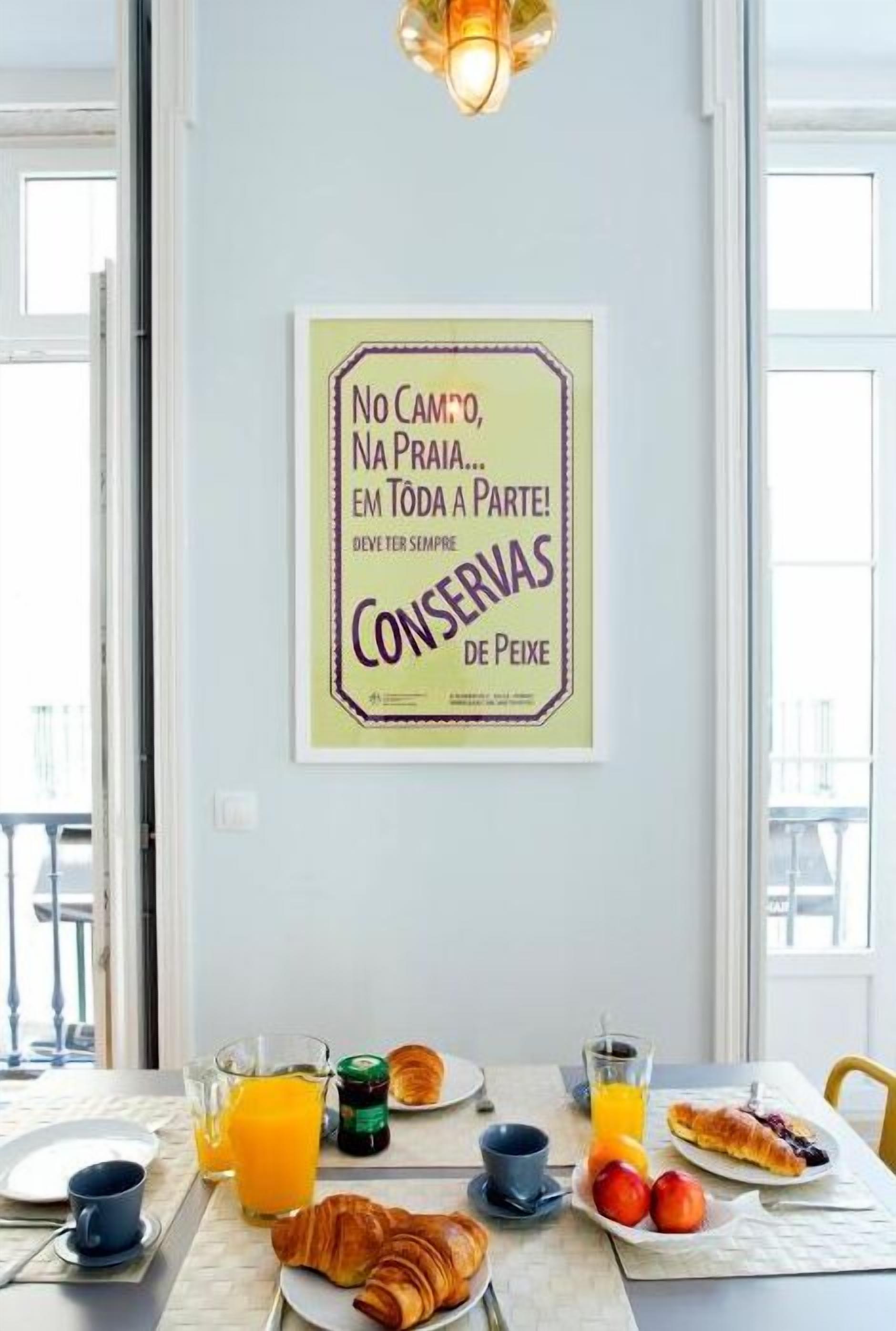 A breakfast table with croissants, tomatoes, orange juice, and a coffee cup. A framed quote is on the wall.