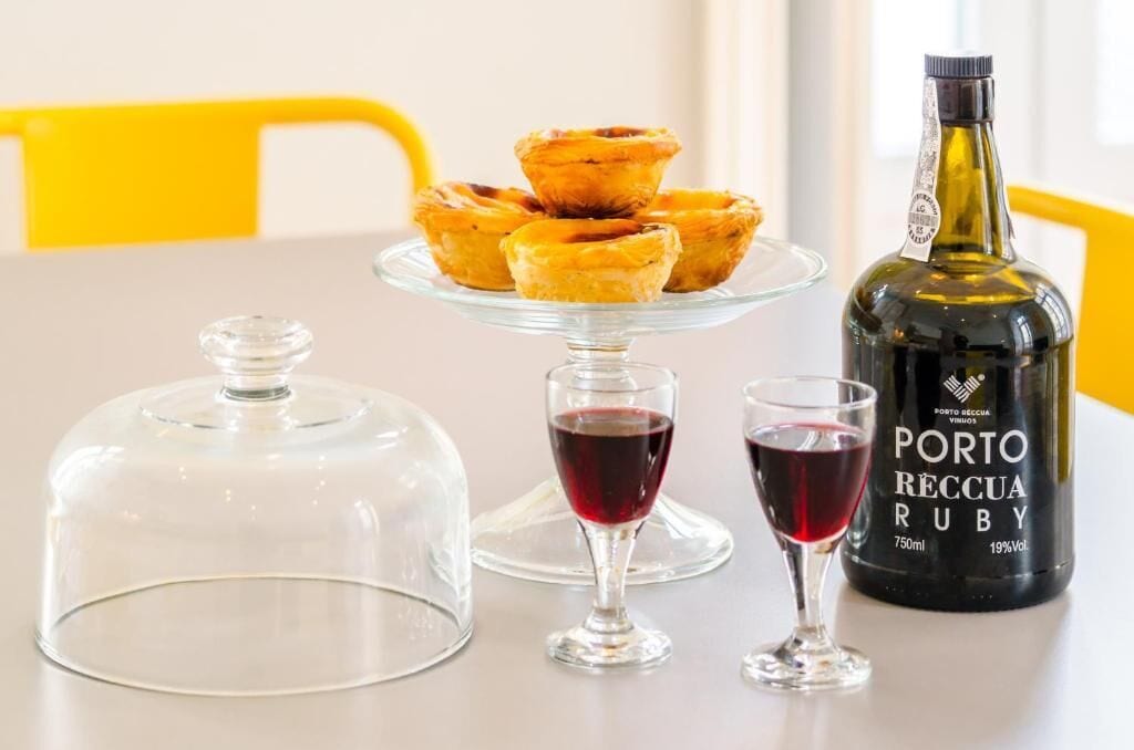 A table with a glass dome cover, two wine glasses, a bottle of Porto Reccia Ruby, and a plate with pastries.