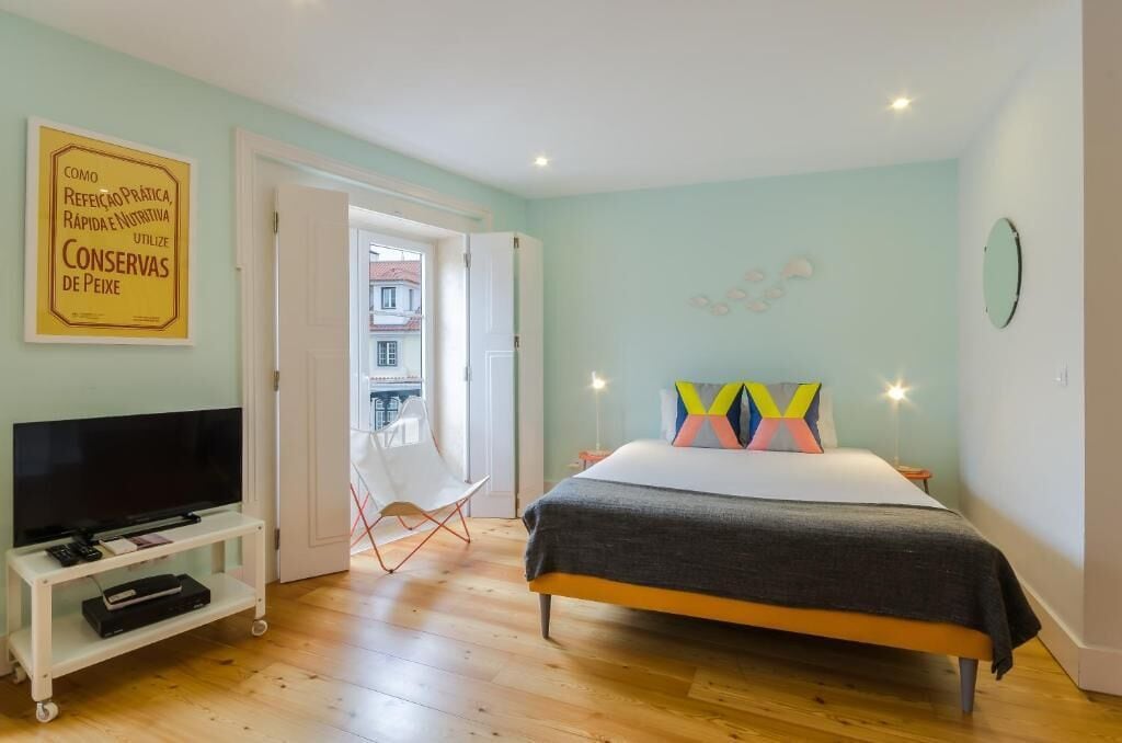 A bedroom with a bed, a TV on a stand, a chair, and a door leading to a balcony.