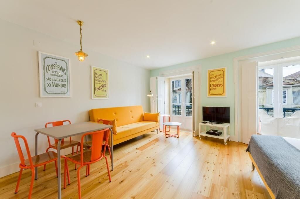 Une pièce avec un plancher en bois, un canapé orange, des fauteuils orange, une petite table blanche et un lit. Il y a deux panneaux encadrés accrochés au mur et un téléviseur posé sur un meuble.