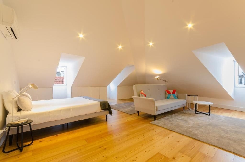 A modern loft bedroom with a loft bed, a sofa, and a small table. The room has wooden flooring and large windows.