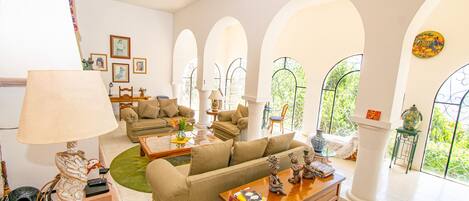 Amplia sala de estar con sofá, mesa de centro y elementos decorativos. Varias ventanas con diseño arqueado ofrecen vistas a zonas verdes.
