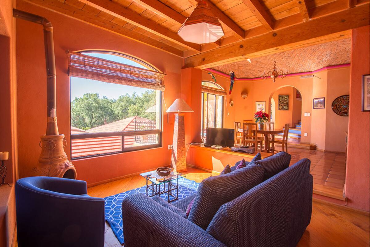 A spacious living room with a large sofa, a television, and a fireplace. There is a dining area with a table and chairs, and a staircase leading to an upper level.