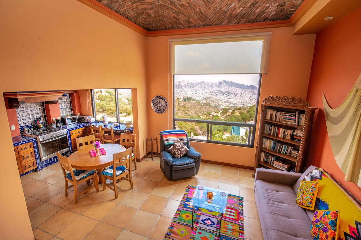 A spacious living area with a dining table, a sofa, a bookshelf, and a large window offering a view of the mountains.