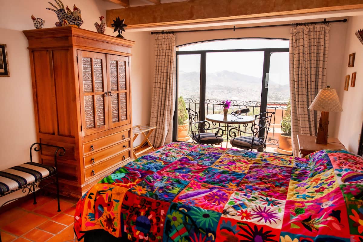 A bedroom with a colorful quilt, wooden dresser, and a view of mountains through a large window.