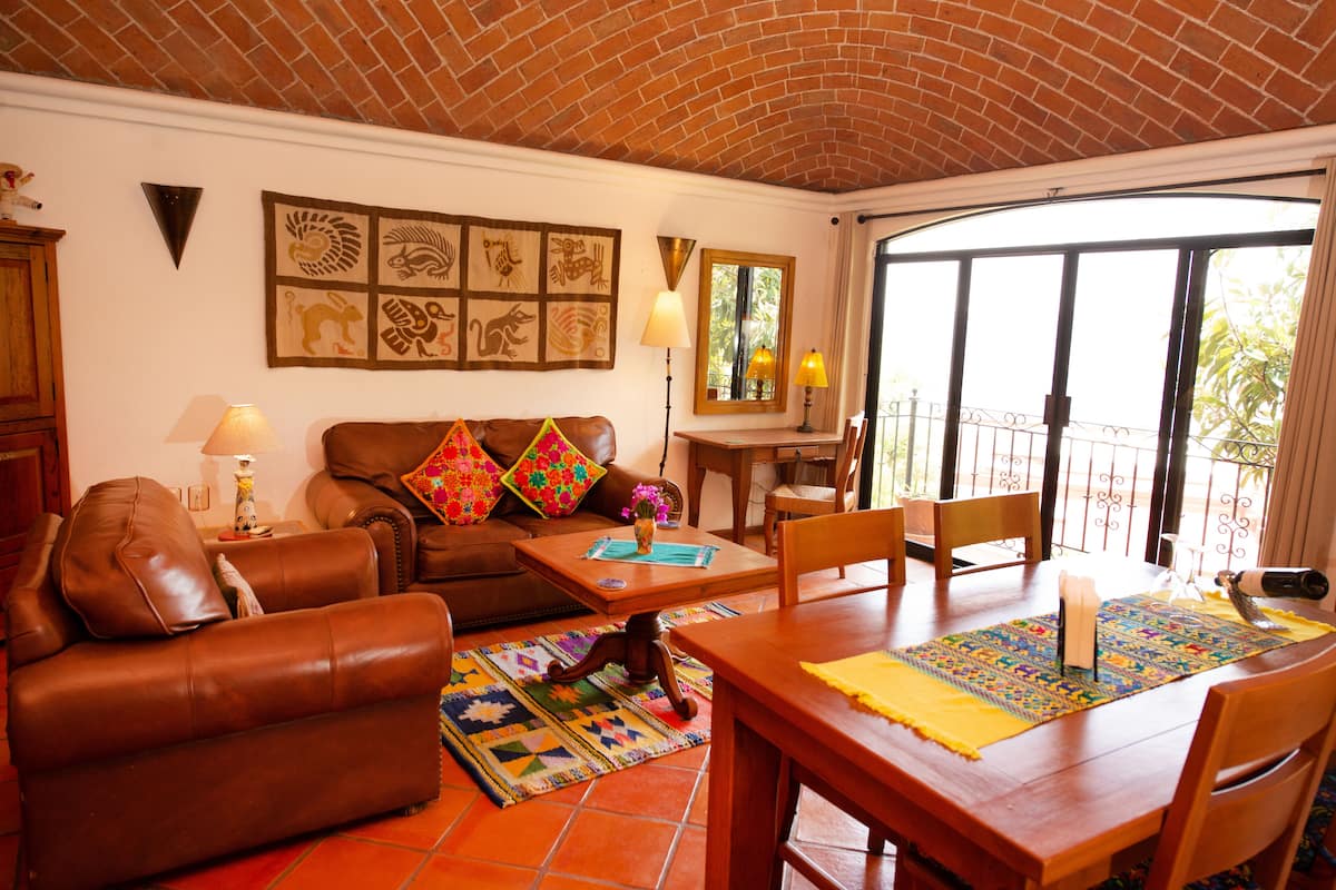 A cozy living room with a brown leather sofa, a wooden coffee table, and a dining table with chairs. There is a balcony with a view of greenery.