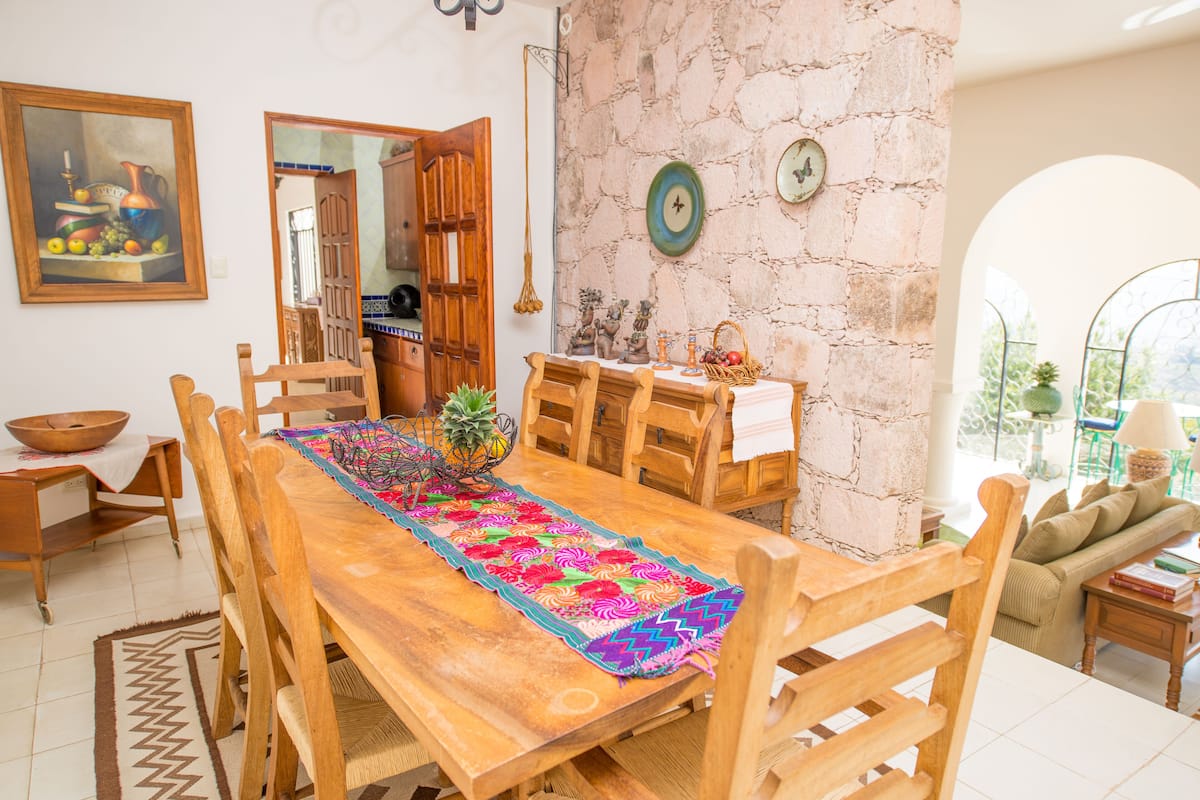 A wooden dining table with chairs, a colorful table runner, and a stone wall with a clock.