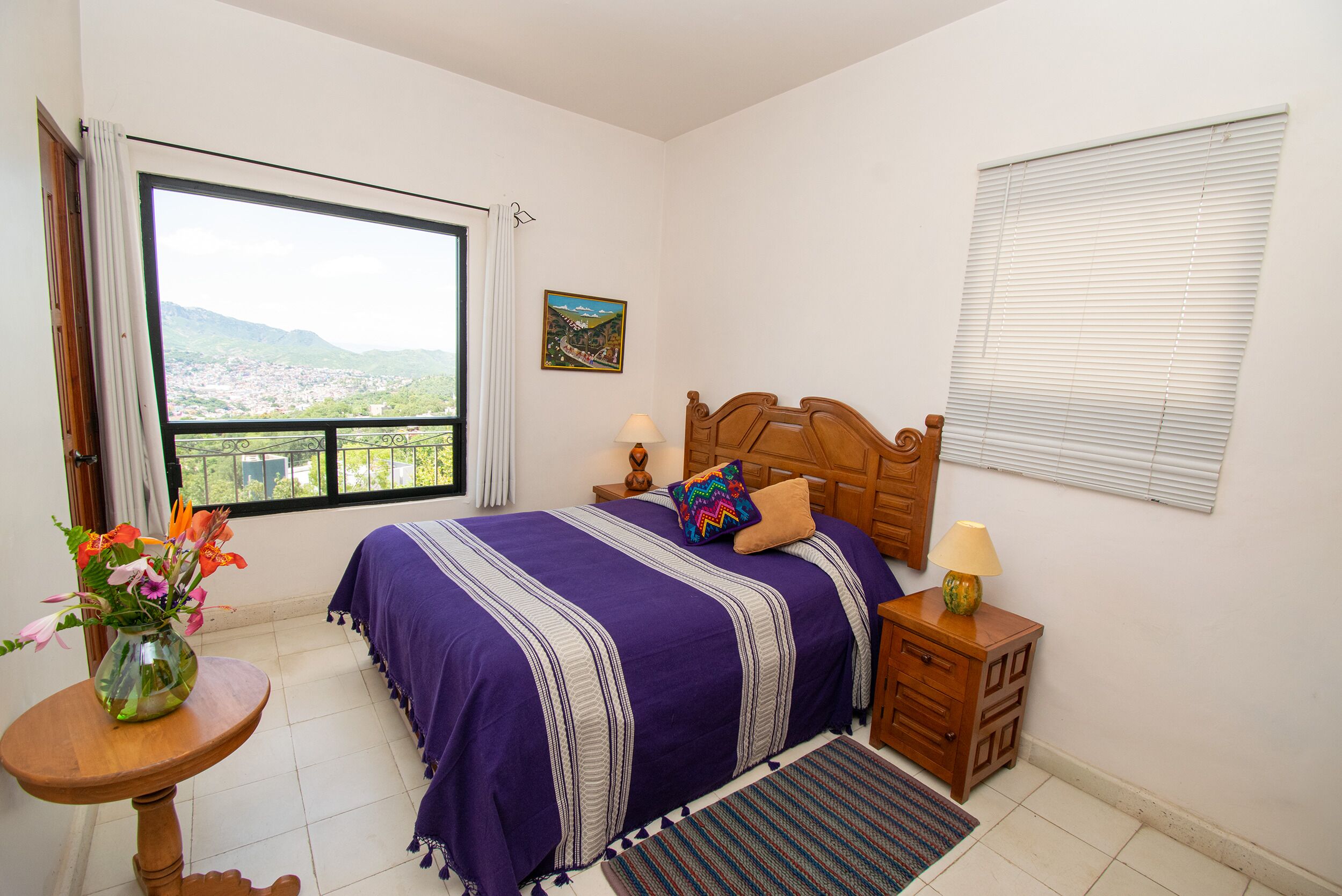 A bedroom with a large window, a bed with a purple bedspread, a nightstand with a lamp, and a small table with a vase of flowers.
