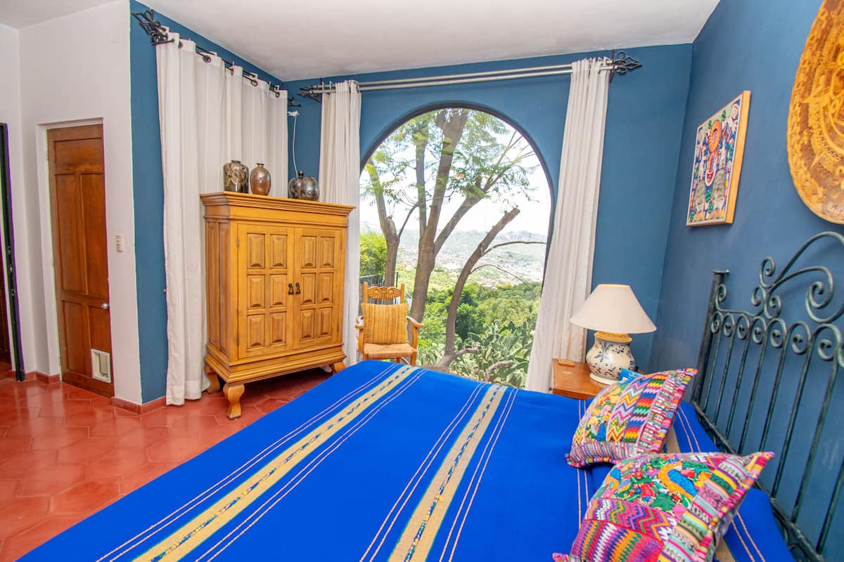 A bedroom with a large bed, a wooden wardrobe, a window with a view of trees, and a colorful wall decoration.