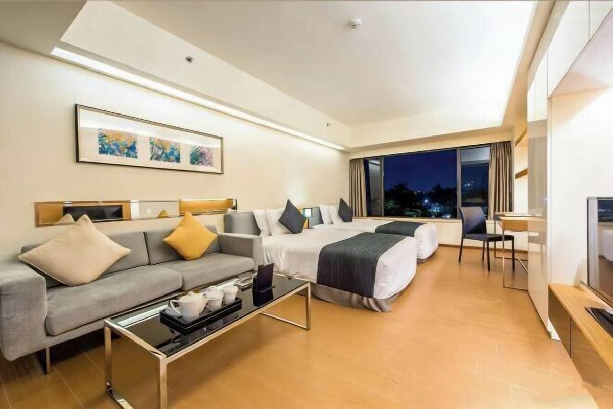 A modern hotel room with a sofa, two single beds, a desk, and a large window with a night view.