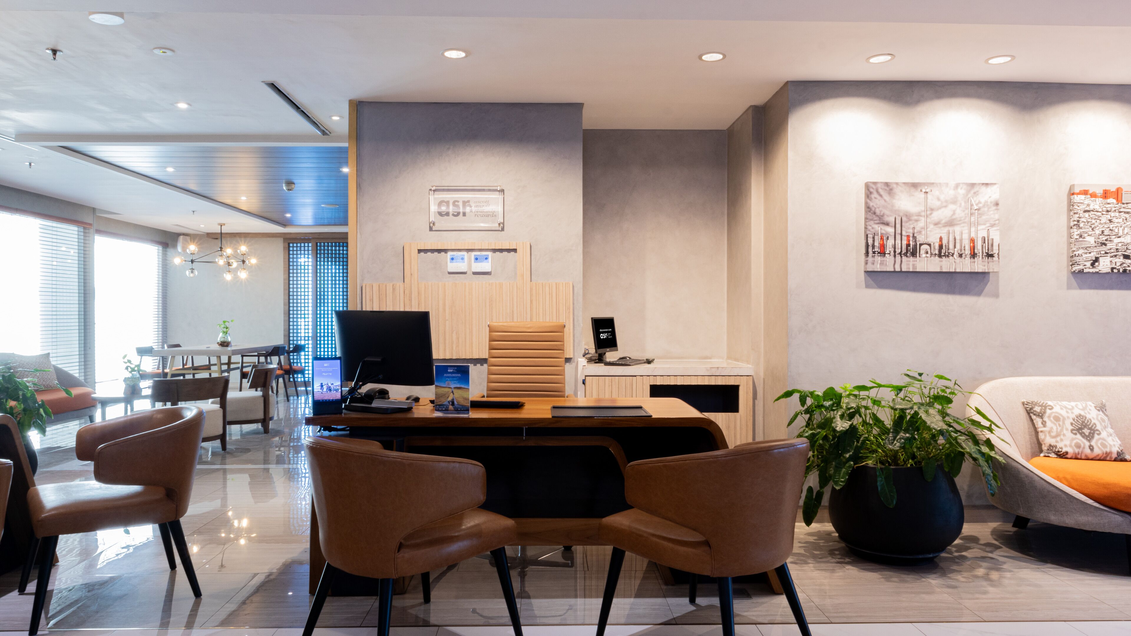 A modern office space with a desk, chairs, a computer, and a sofa. There are plants and artwork on the walls.