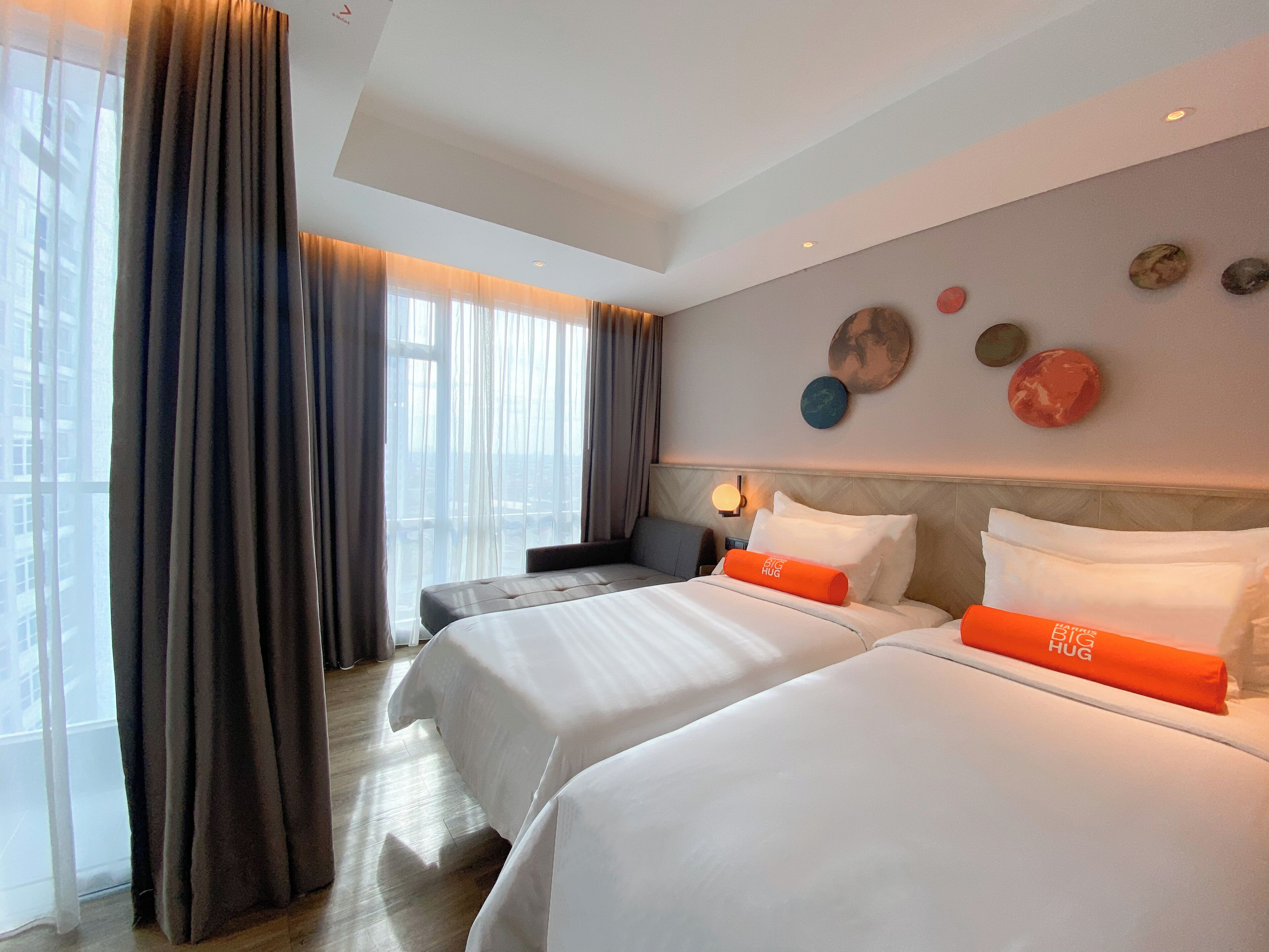 A hotel room with two beds, a large window with curtains, a wall with decorative plates, and a nightstand with a lamp.