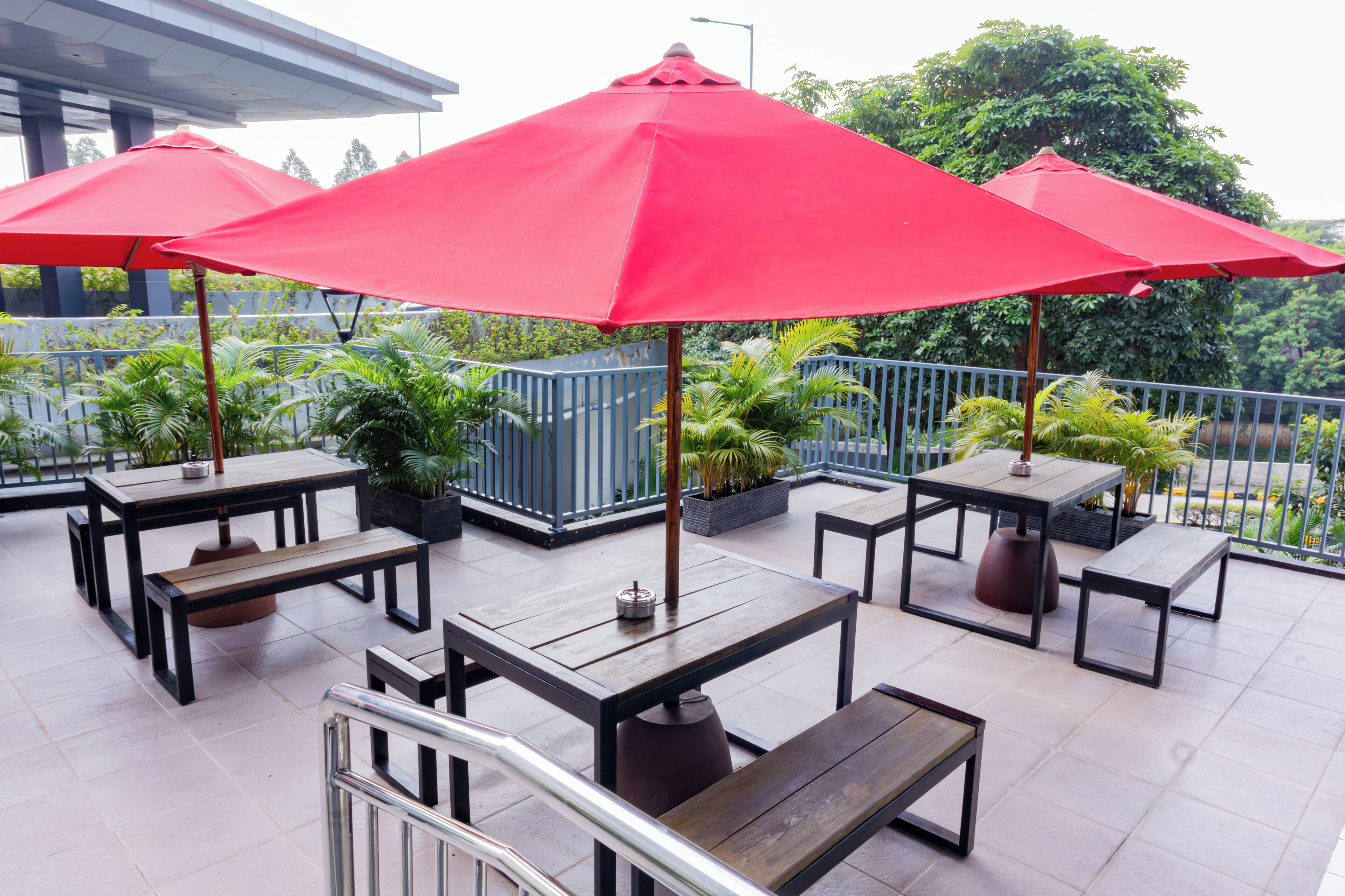 Een terras buiten met tafels en banken, rode parasols en potplanten.