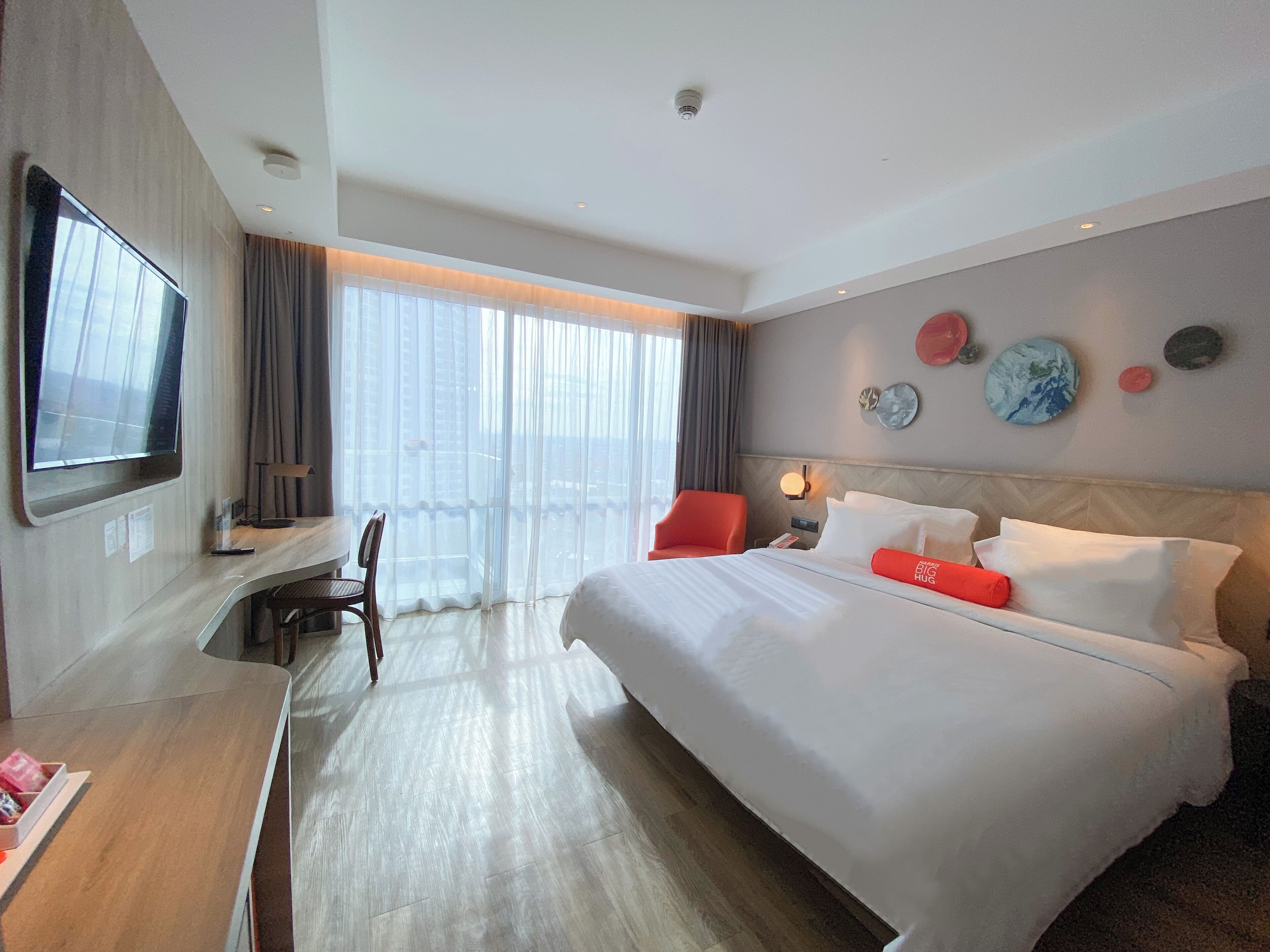 A hotel room with a large bed, a desk with a chair, a TV, and a view of the city through large windows.