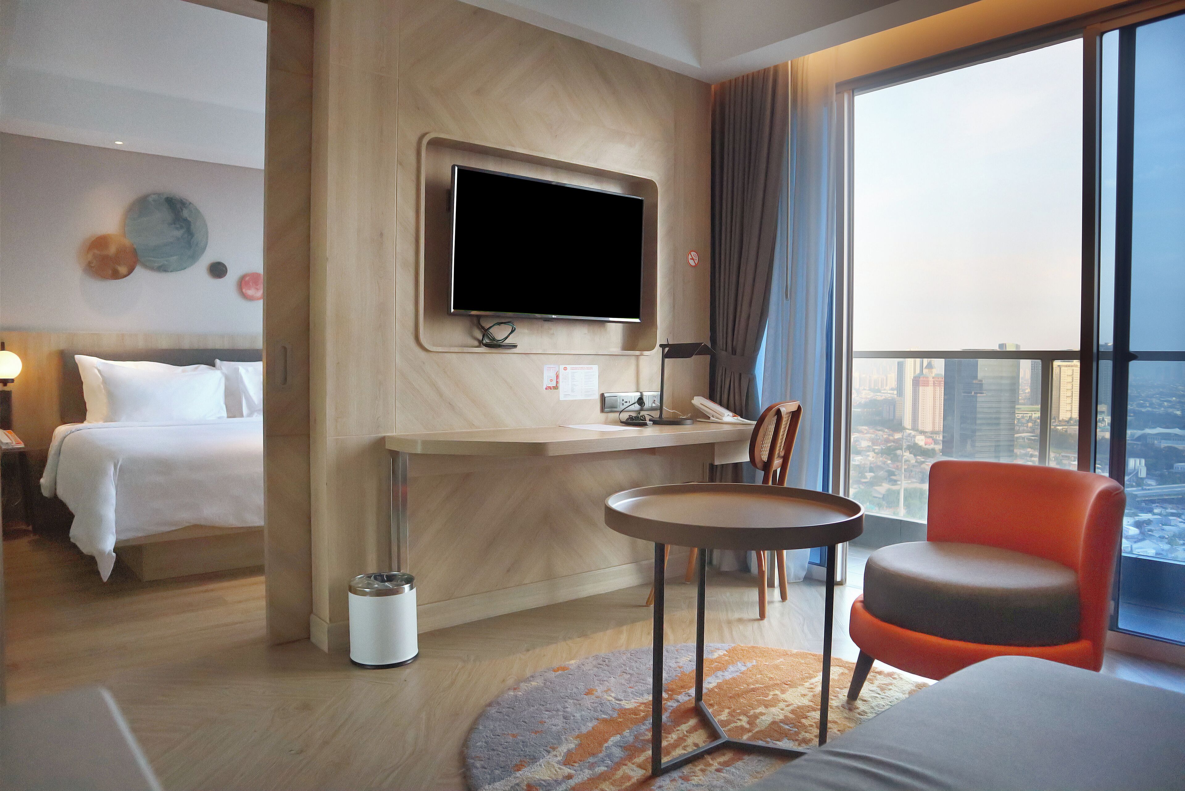 A hotel room with a bed, a desk, a chair, a television, and a large window with a city view.