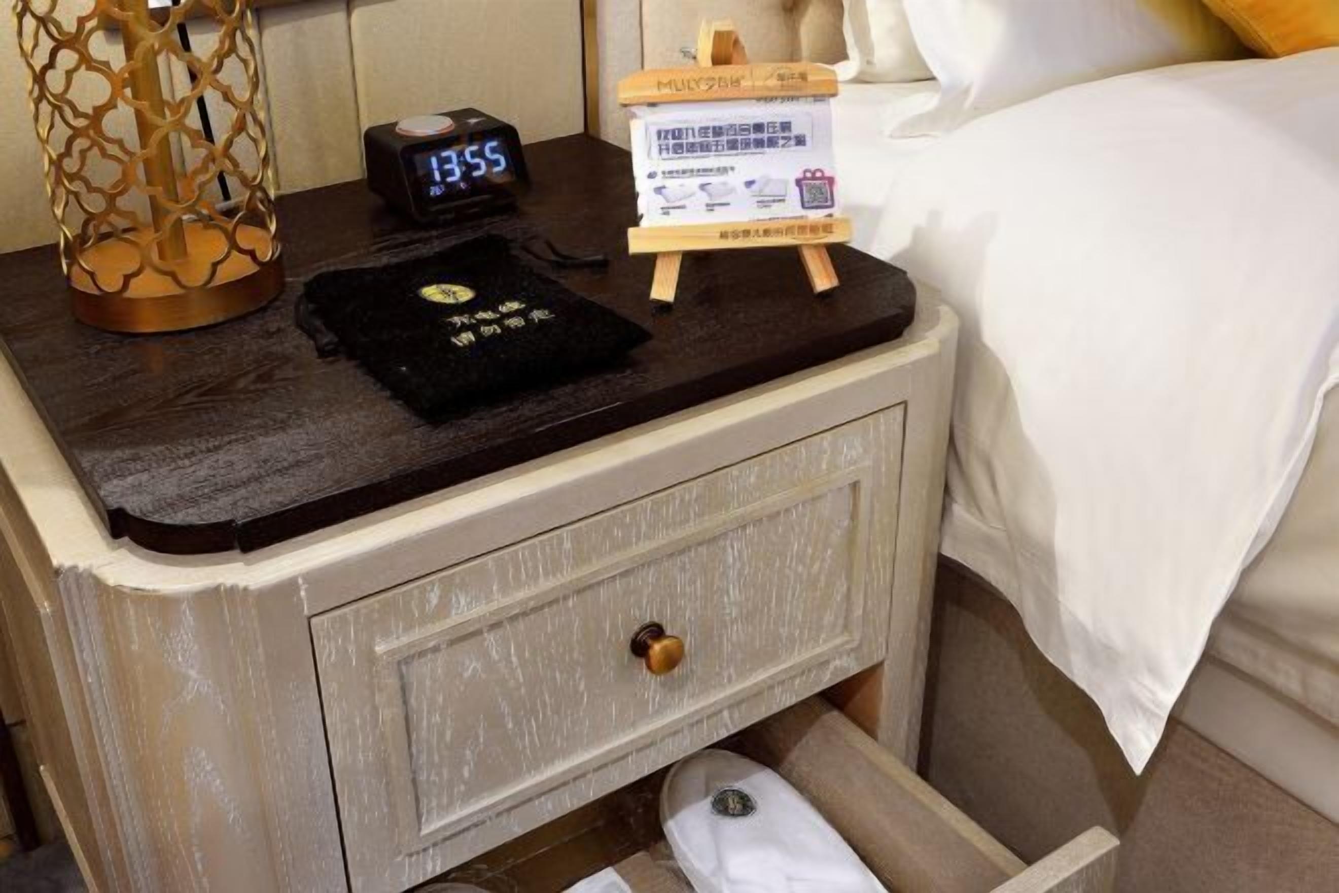 A wooden nightstand with a black clock, a small wooden stand with a sign, and a drawer with a white towel.