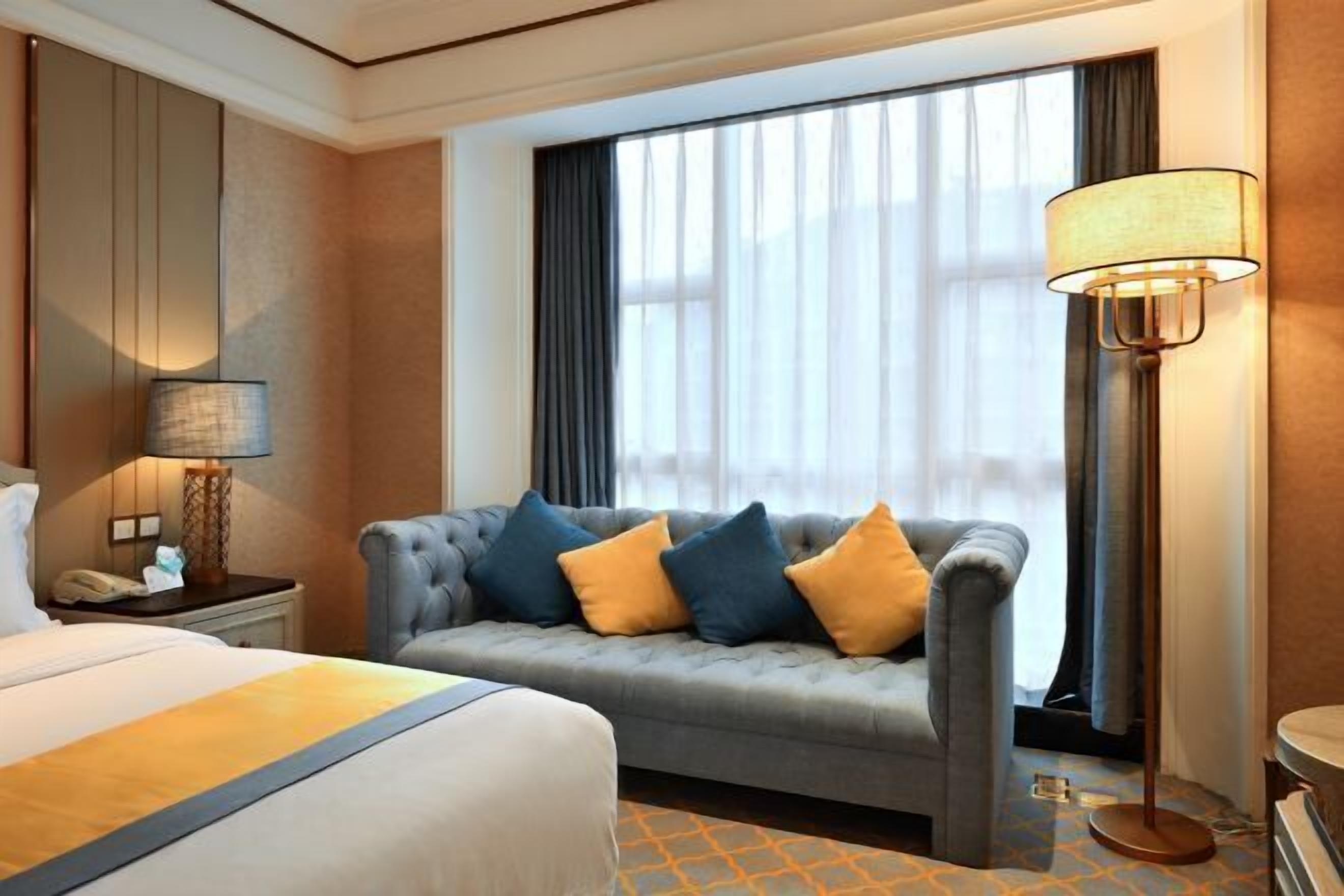 A hotel room with a bed, a grey sofa with blue and yellow cushions, a standing lamp, and a large window with sheer curtains.