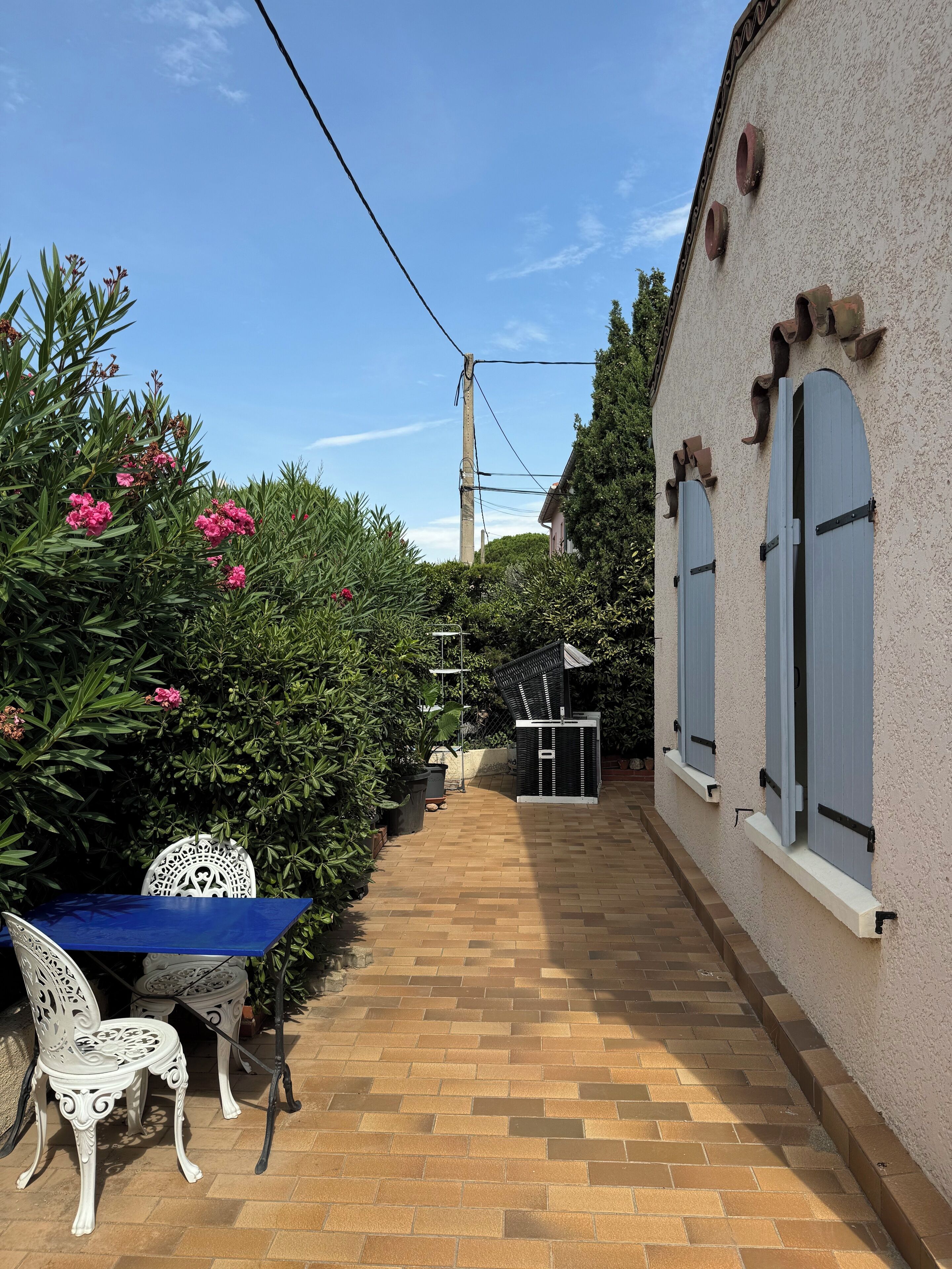 Un vialetto in mattoni che conduce a un edificio bianco con persiane blu, immerso nel verde e fioriture.