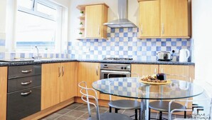 A modern kitchen with wooden cabinets, a gas stove, a stainless steel range hood, and a glass dining table with chairs. - Beautiful 5-bed House Roker Beach (Sunderland)