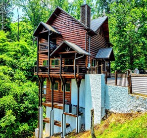 A multi-level log cabin with a balcony, surrounded by lush greenery. - Whiskey Bear Creek: 2021, SAUNA, JACUZZI, AND POOL access! (Gatlinburg)