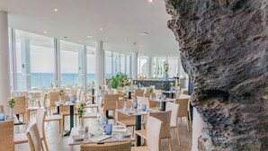 Restaurante com mesas preparadas para refeições, grandes janelas com vista para o mar e uma parede de pedra.