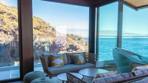 Interior moderno com sofá, cadeiras e mesa, com vista para a costa rochosa e o mar.