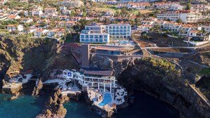 Vista aérea de um hotel à beira-mar, com vários edifícios, piscina e costa rochosa.