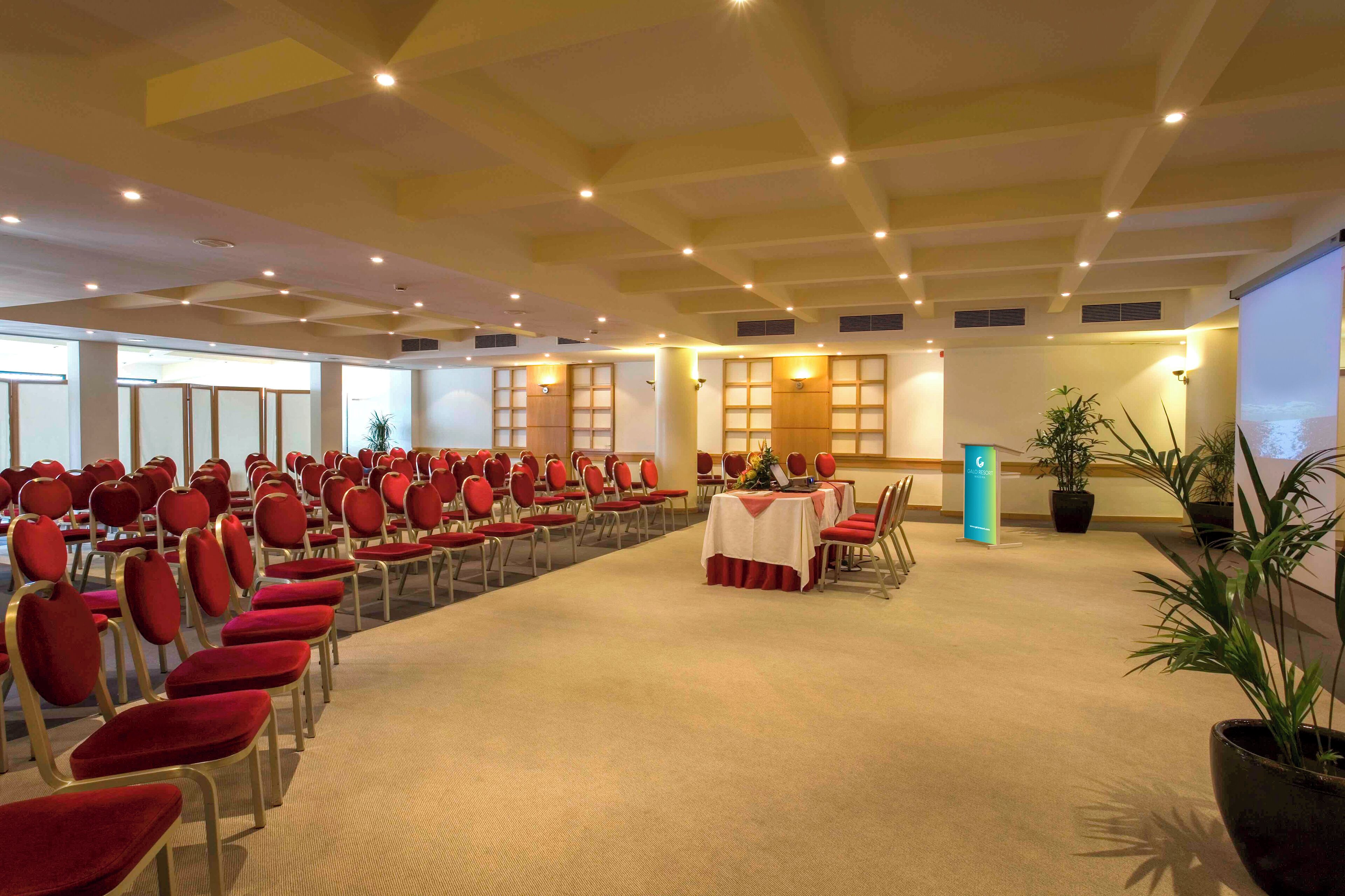 Une salle de conférence avec des rangées de chaises et de tables rouges, une scène avec un pupitre et des plantes en pot.
