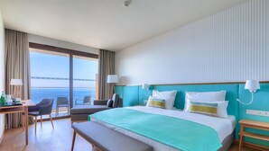 Quarto de hotel com uma cama grande, uma escrivaninha, uma cadeira, um banco e vista para o mar.