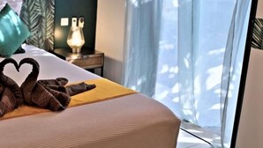 Une chambre d’hôtel avec un lit, une décoration en forme de cœur réalisée avec des serviettes, une table de chevet avec une lampe et une vue sur l’extérieur à travers des rideaux transparents.